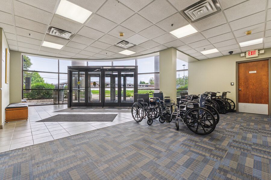 Spacious building entrance with glass double doors, gray carpet, drop ceiling, and multiple wheelchairs parked near the exit.