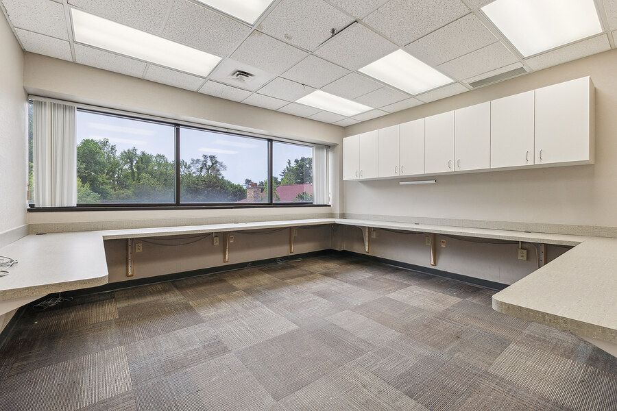 Empty office space with built-in white cabinets, L-shaped desk, large window, and patterned carpet flooring.