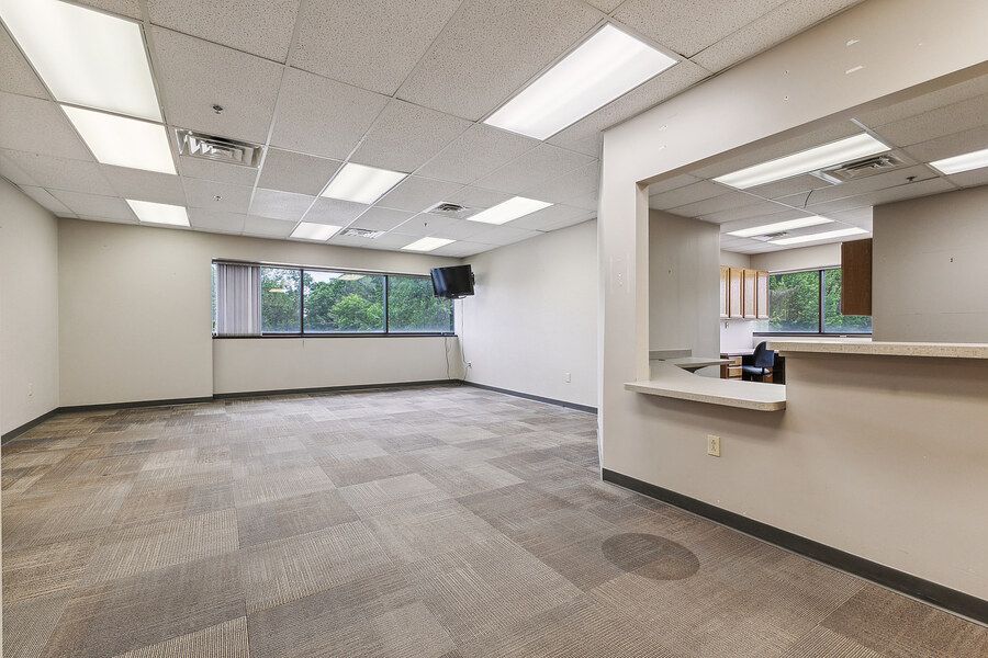Spacious open office space with patterned carpet, large window, and a reception counter cutout in the wall.