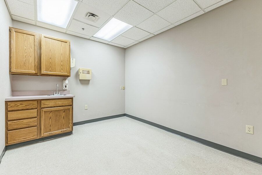 Small medical exam room with oak cabinets, built-in sink, drop ceiling, and neutral carpeted flooring.