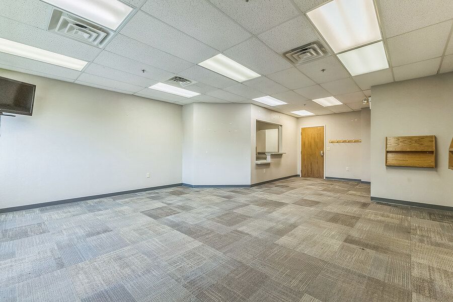Open commercial office space with patterned carpet, drop ceiling, wooden door, and wall-mounted bulletin board.