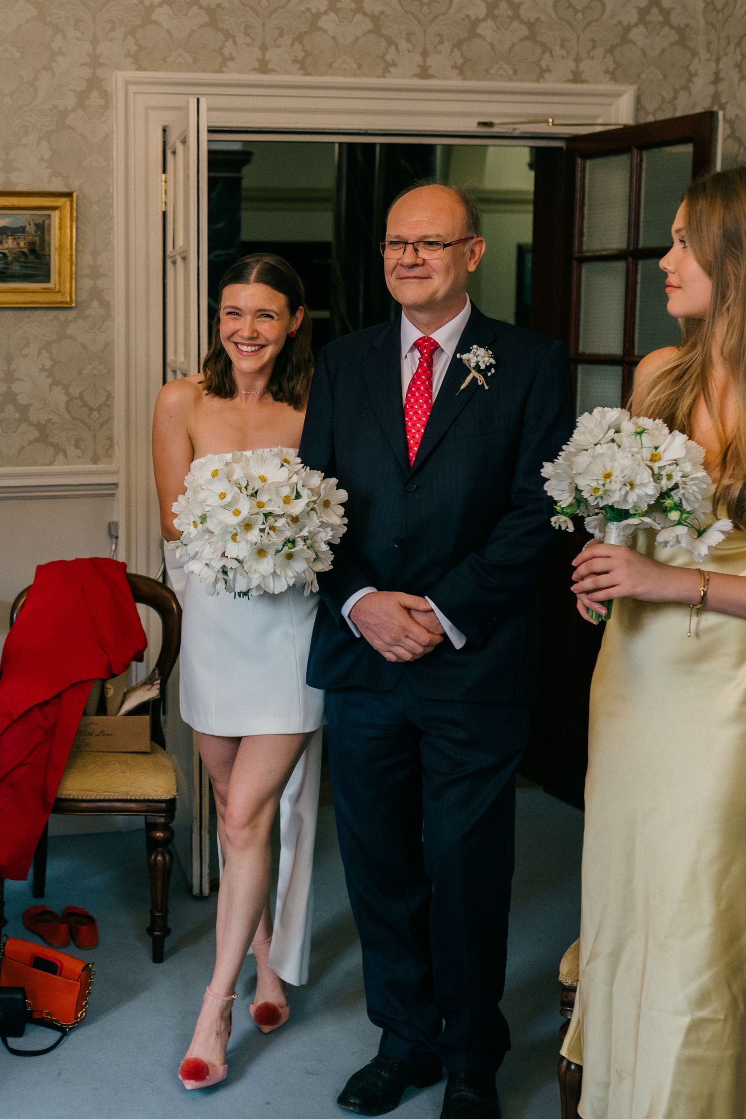 Bride in white mini dress holds white bouquet beside suited man with red tie and bridesmaid in yellow dress.