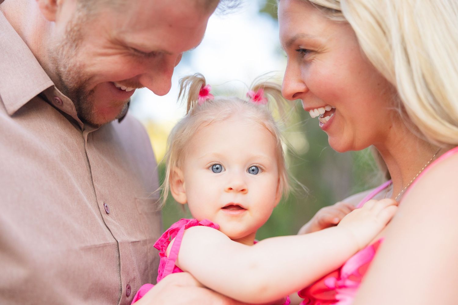 Smiling parents lovingly gazing at their blue-eyed toddler girl outdoors.