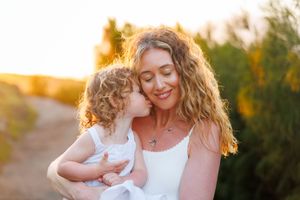 A curly-haired toddler gives a tender kiss on the cheek to a smiling woman holding her at sunset.