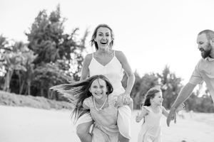 Black and white photo of a laughing mother giving her daughter a piggyback ride on a sandy beach.