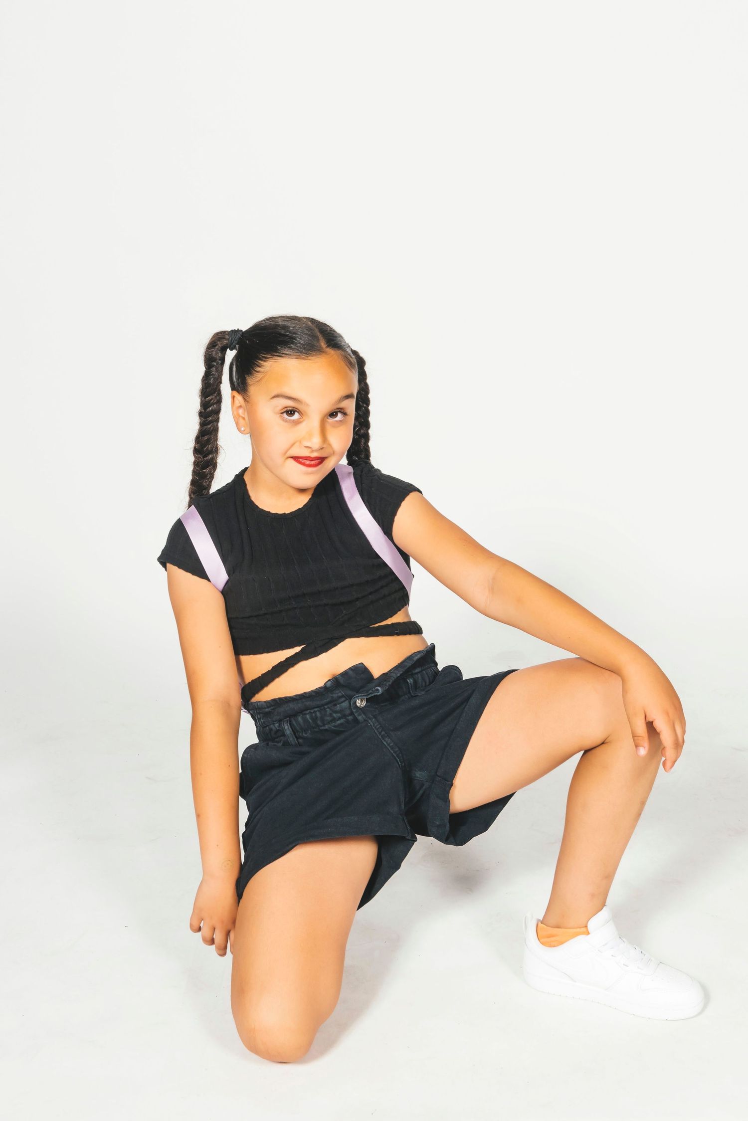 Young girl in black crop top and shorts with braids, posing on one knee against white background.