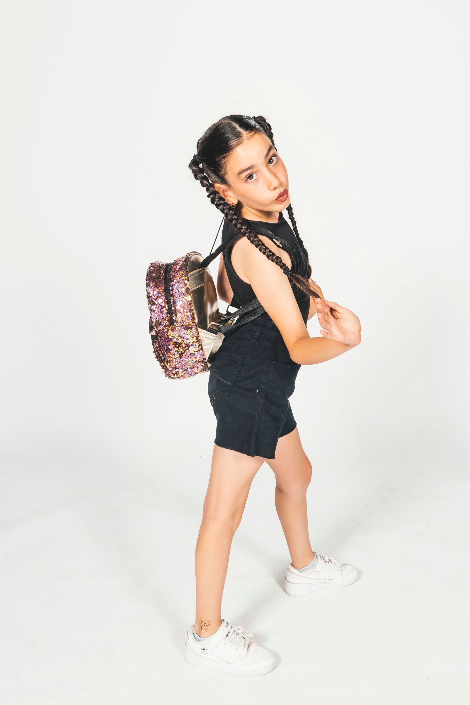 Young girl with braided hair wearing black outfit and sparkly pink sequin backpack, posing on white background.