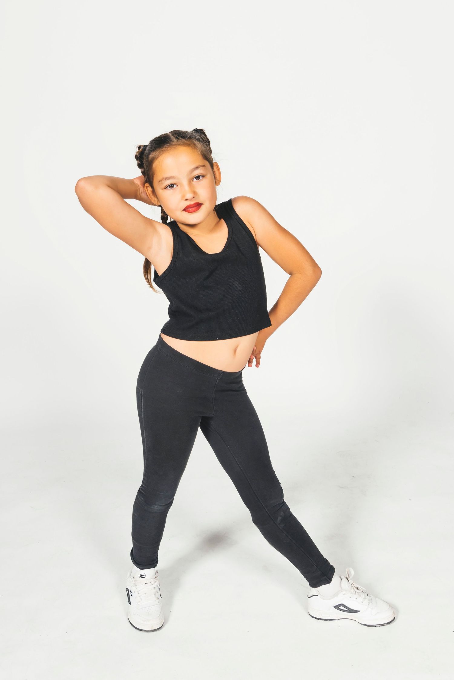 Young girl in black crop top and leggings poses confidently in white sneakers against a white background.
