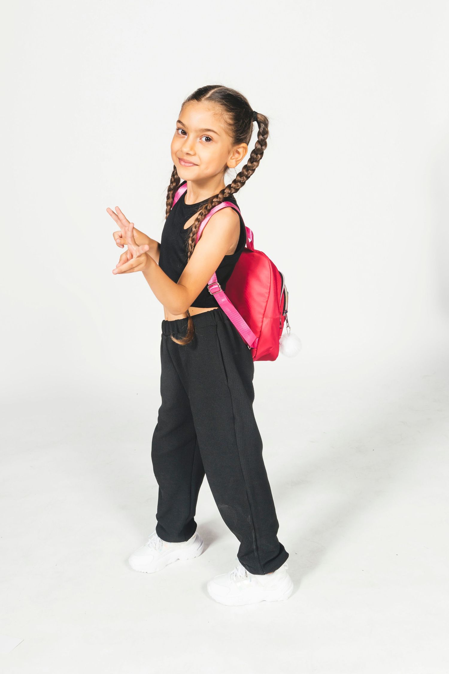 Young girl in black outfit with pink backpack making peace sign, studio photo.