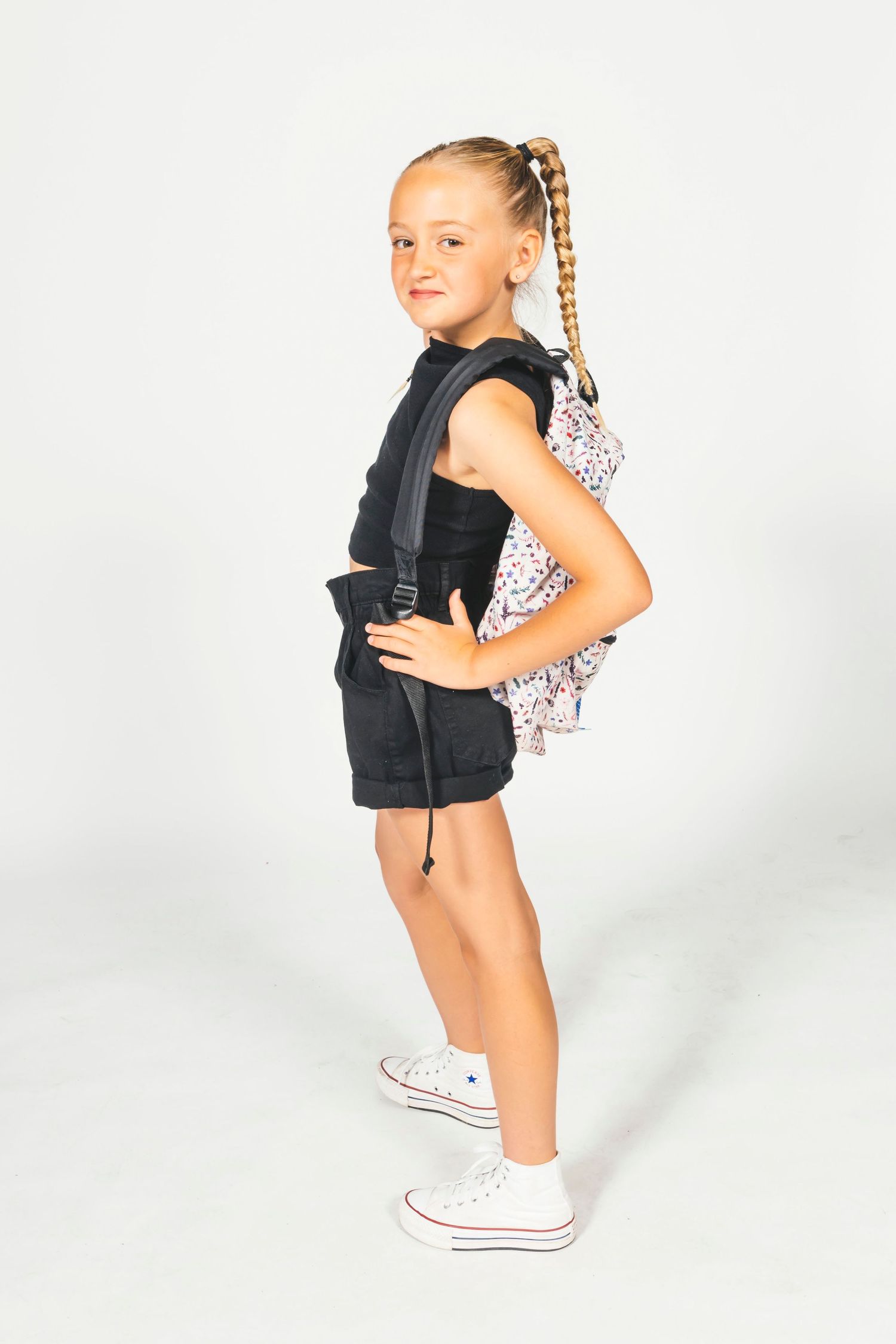 Blonde girl in black outfit with floral backpack posing in white studio.