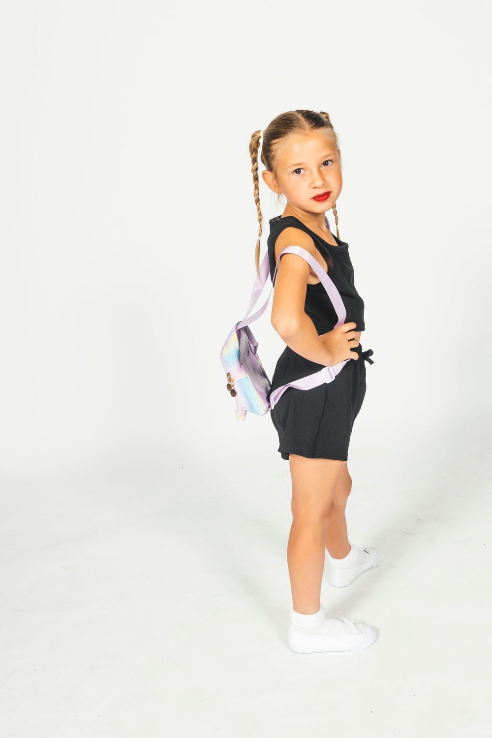 Young girl in black romper with braided pigtails and colorful backpack posing against white background.