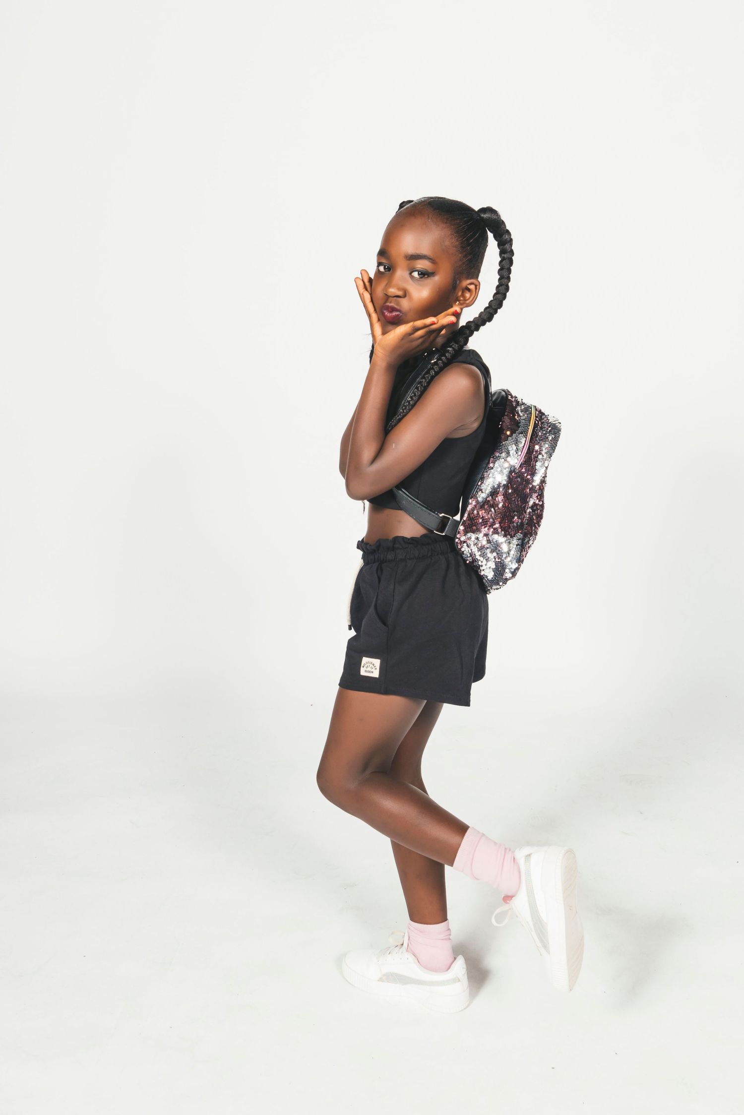 Young Black girl in black athletic outfit with patterned backpack poses confidently against white background.