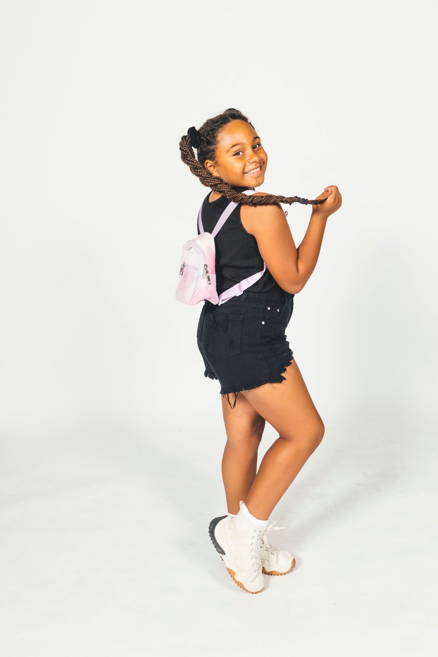 Young girl in black romper with pink mini backpack, posing playfully with braided hair on white background.