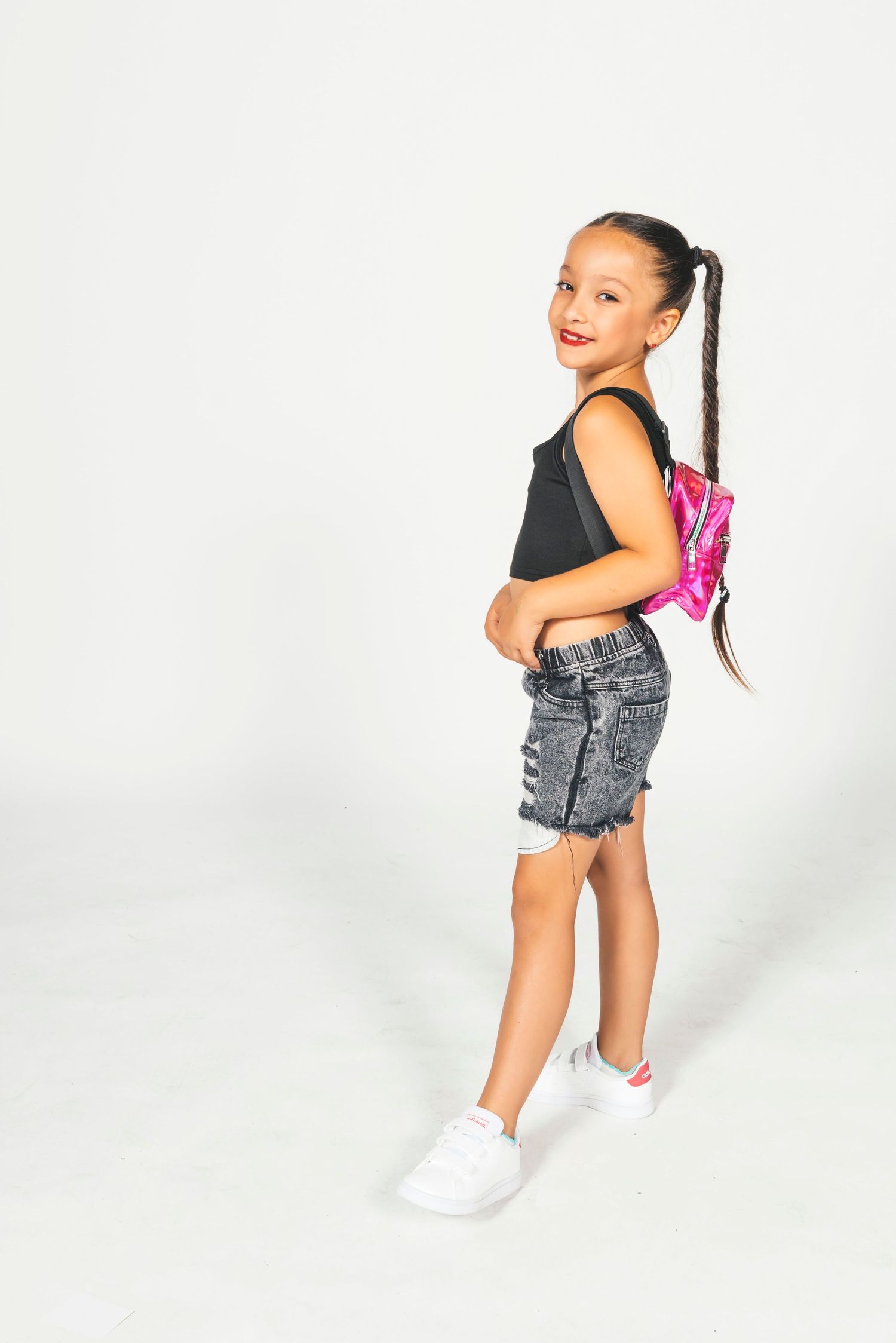 Young girl in black top, denim shorts, white sneakers, and pink fanny pack poses against white background.
