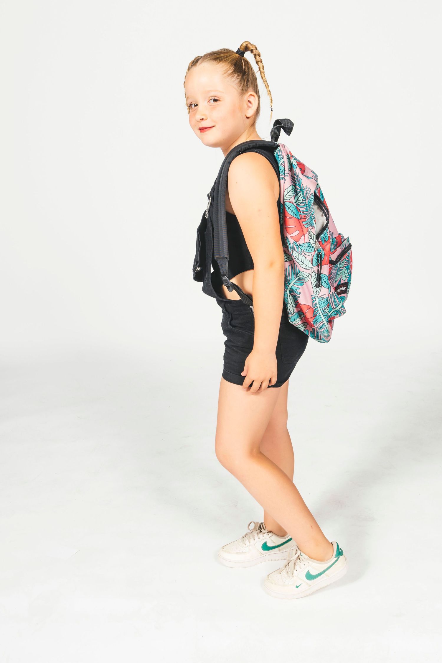 Young girl with pigtail wearing black outfit and colorful backpack, posing against white background.