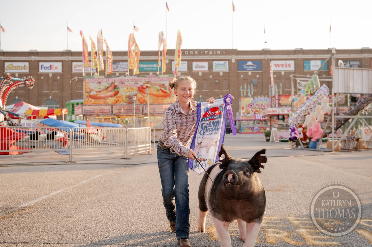 Two children lead pigs down a fairground path at York Fair, with colorful carnival games and a Weis store in the background.