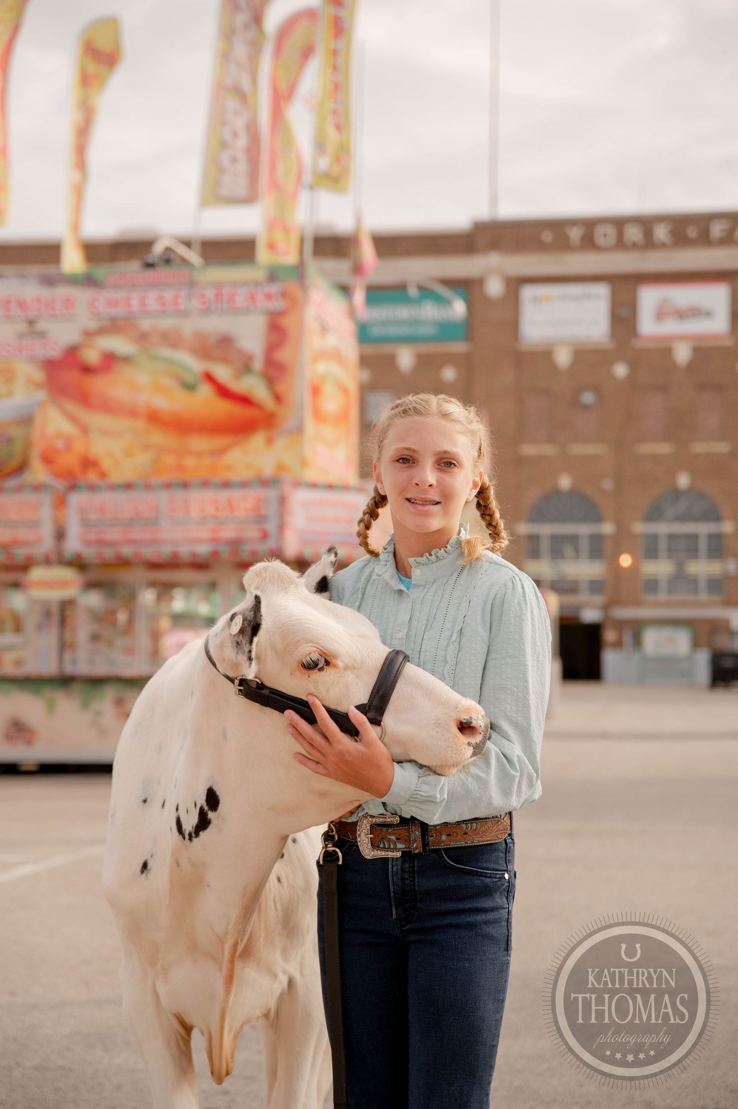 Young girl with braids holds a white spotted calf at a county fair with colorful food stands behind her.