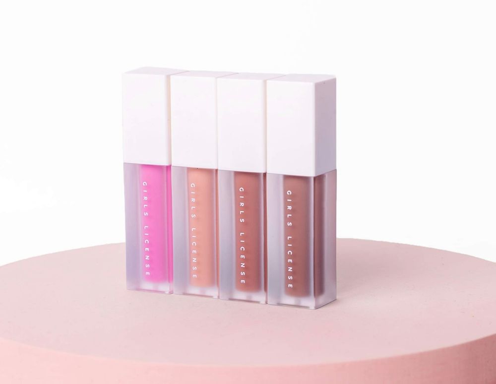 Four lip gloss tubes in pink, peach, nude, and mauve shades displayed on a pink pedestal against a white background.