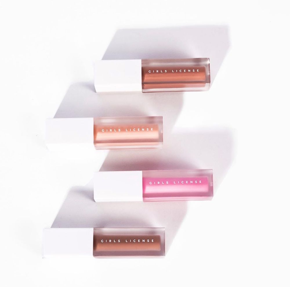 Three Girls License lip glosses in bronze, pink, and mauve shades arranged diagonally on a white background.