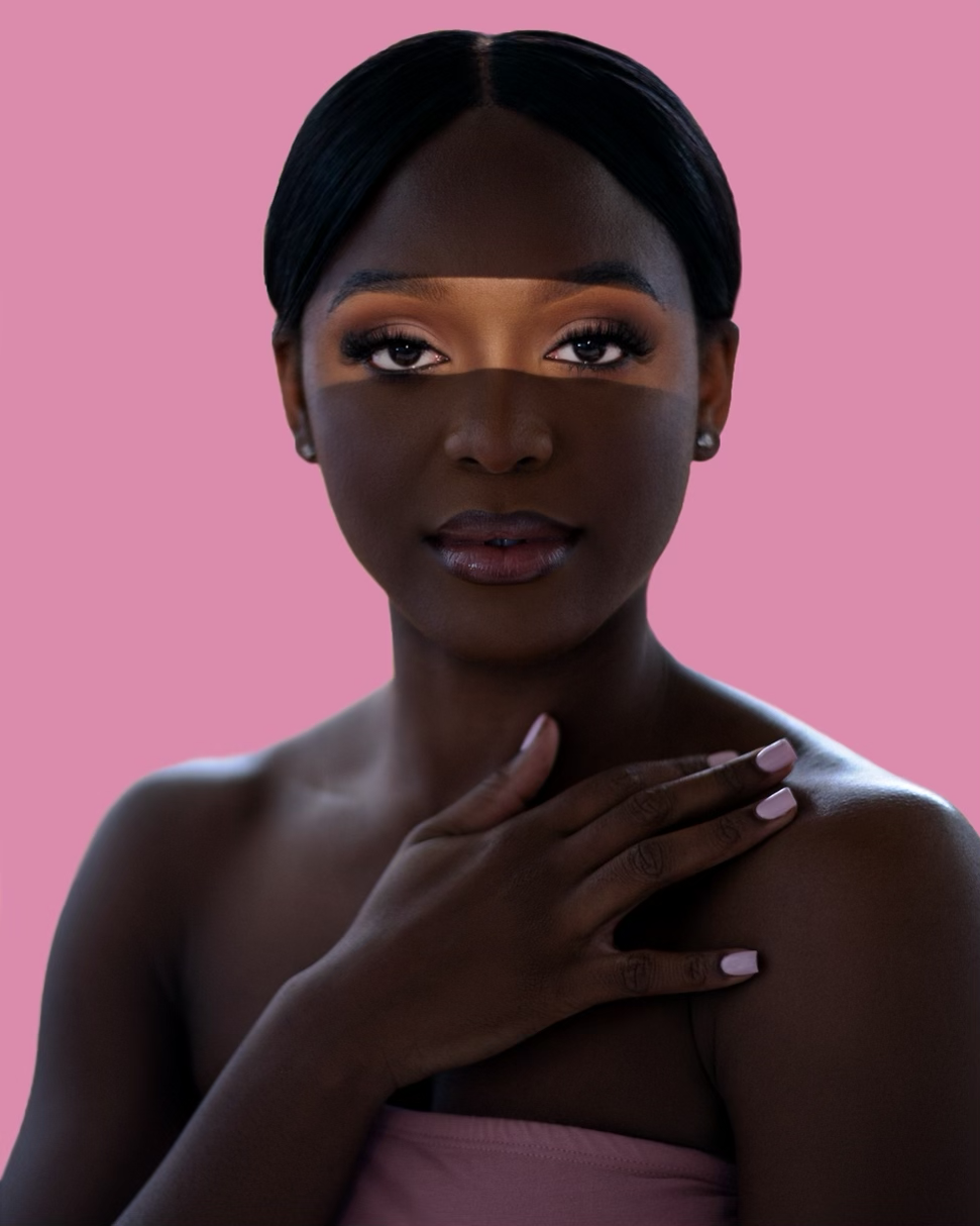 Elegant Black woman with glam makeup touches her neck gracefully against a soft pink studio background.