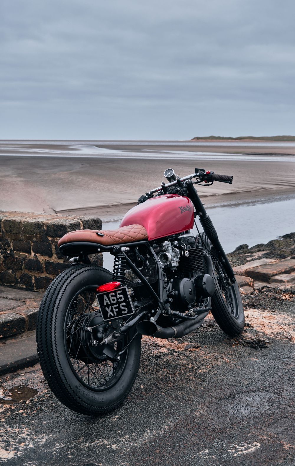 Red custom cafe racer motorcycle parked by a coastal beach on a cloudy day, featuring brown seat and black spoked wheels.