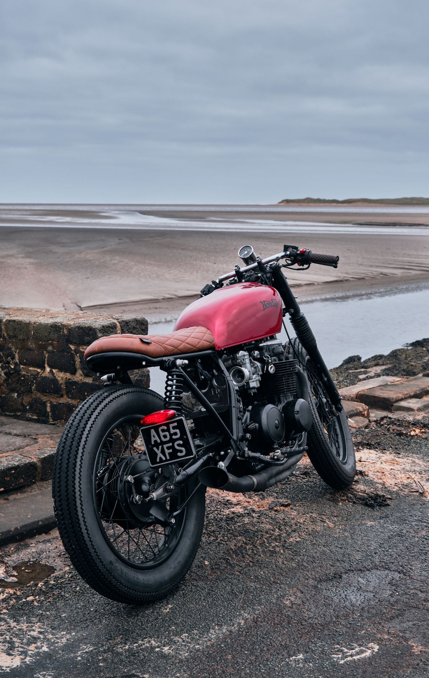 Red custom cafe racer motorcycle parked by a coastal beach on a cloudy day, featuring brown seat and black spoked wheels.
