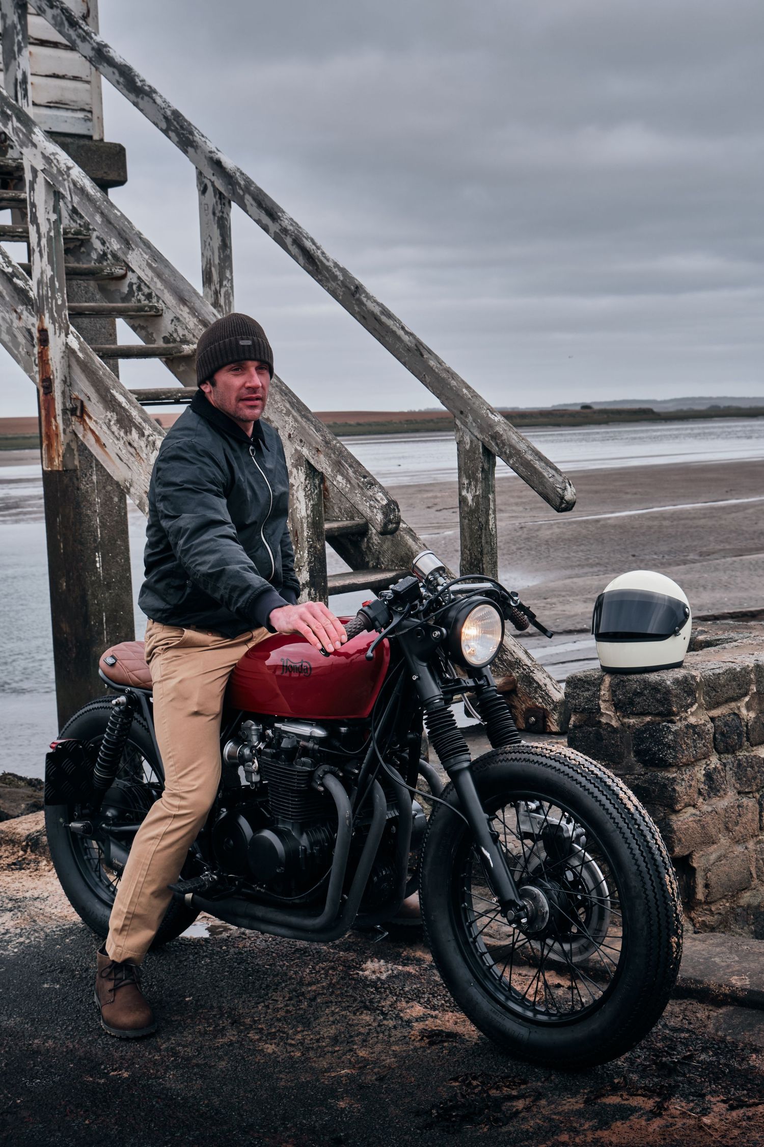 Man in black jacket leaning on a red vintage motorcycle near weathered wooden stairs by a beach.