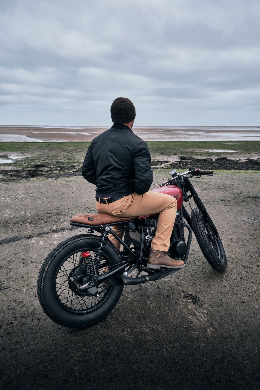 Man in black jacket sitting on vintage cafe racer motorcycle overlooking a coastal landscape under cloudy skies.