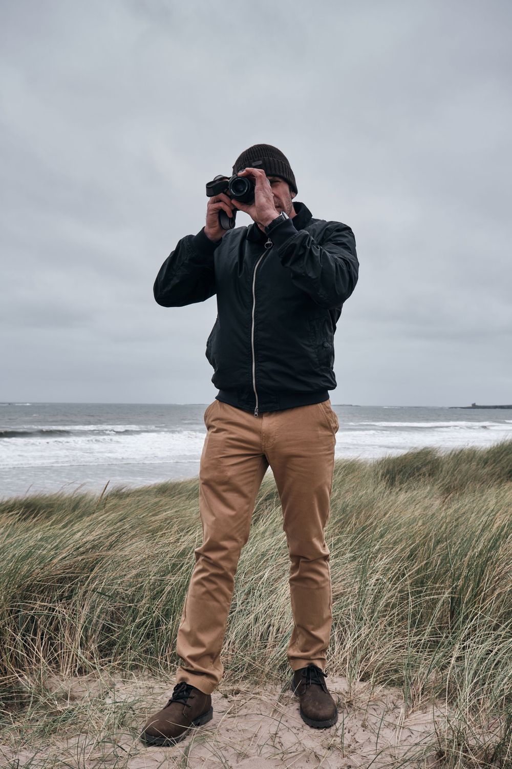 Man in black jacket and tan pants photographing at a beach with dramatic cloudy skies and ocean waves.