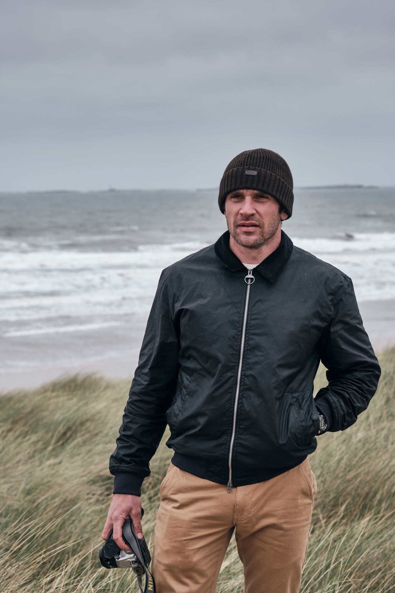 Man in black bomber jacket and beanie hat standing on a windy beach with rough seas in the background.