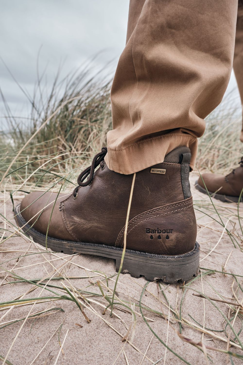 Close-up of brown Barbour leather lace-up boots worn with tan trousers on sandy ground with dry grass.