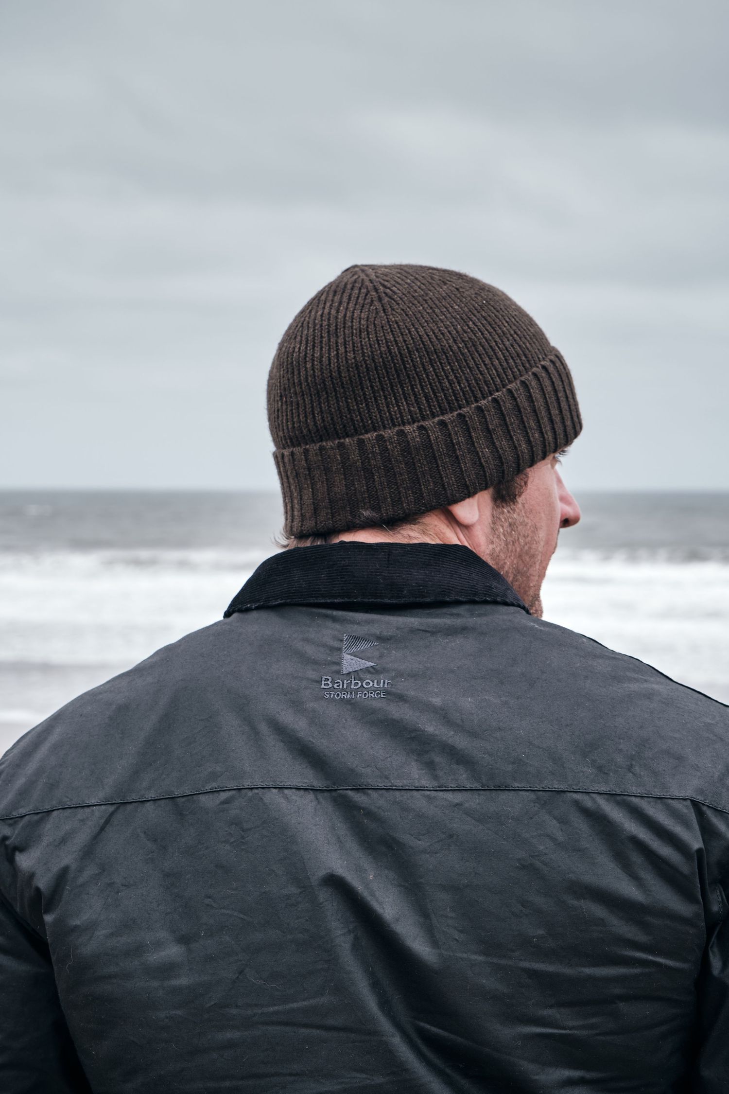 Man in dark Barbour jacket and brown knit beanie hat gazing at stormy ocean waves on overcast day.