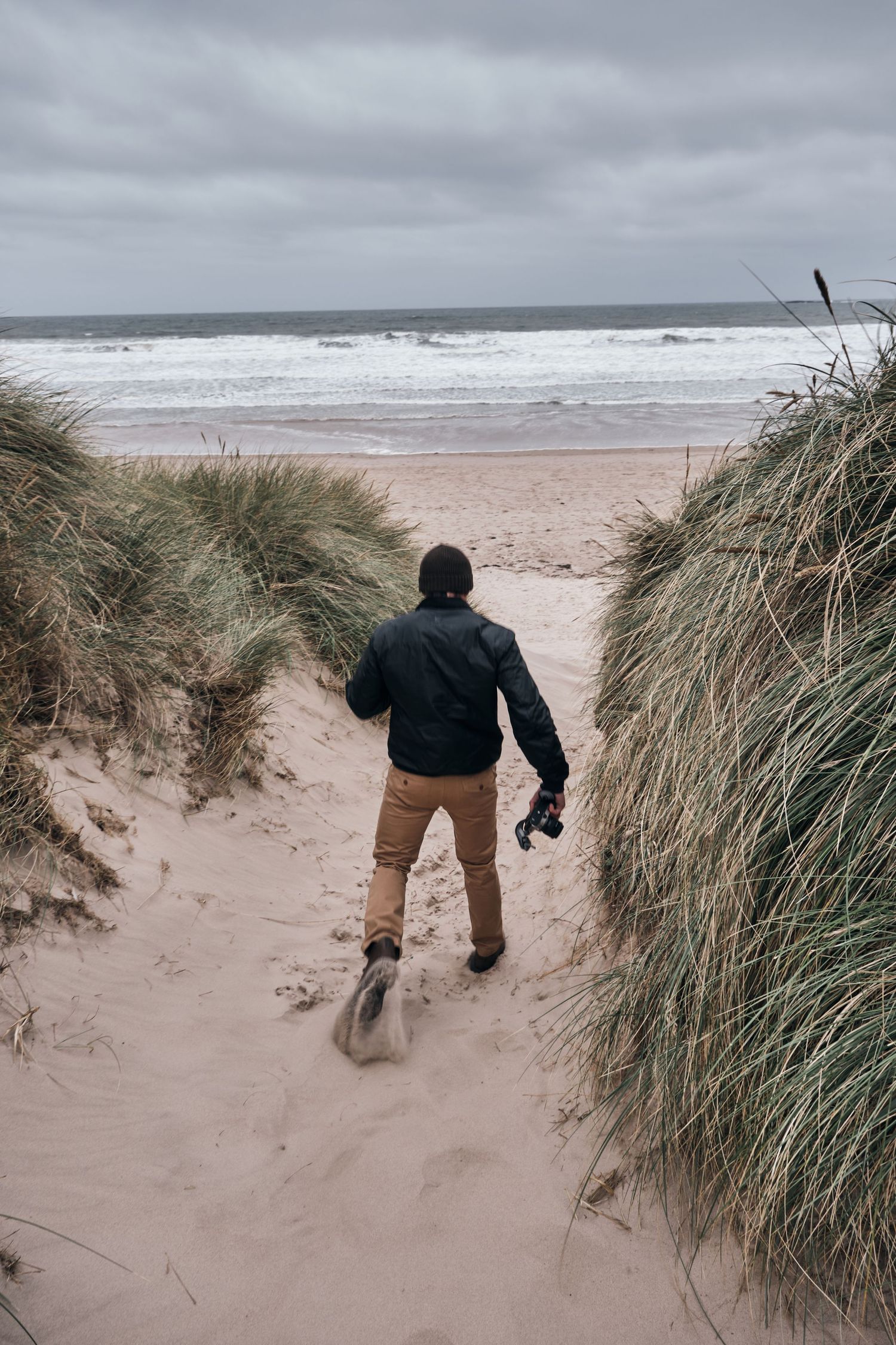 Photographer walking through sand dunes toward a stormy beach, holding camera, wearing black jacket and tan pants.