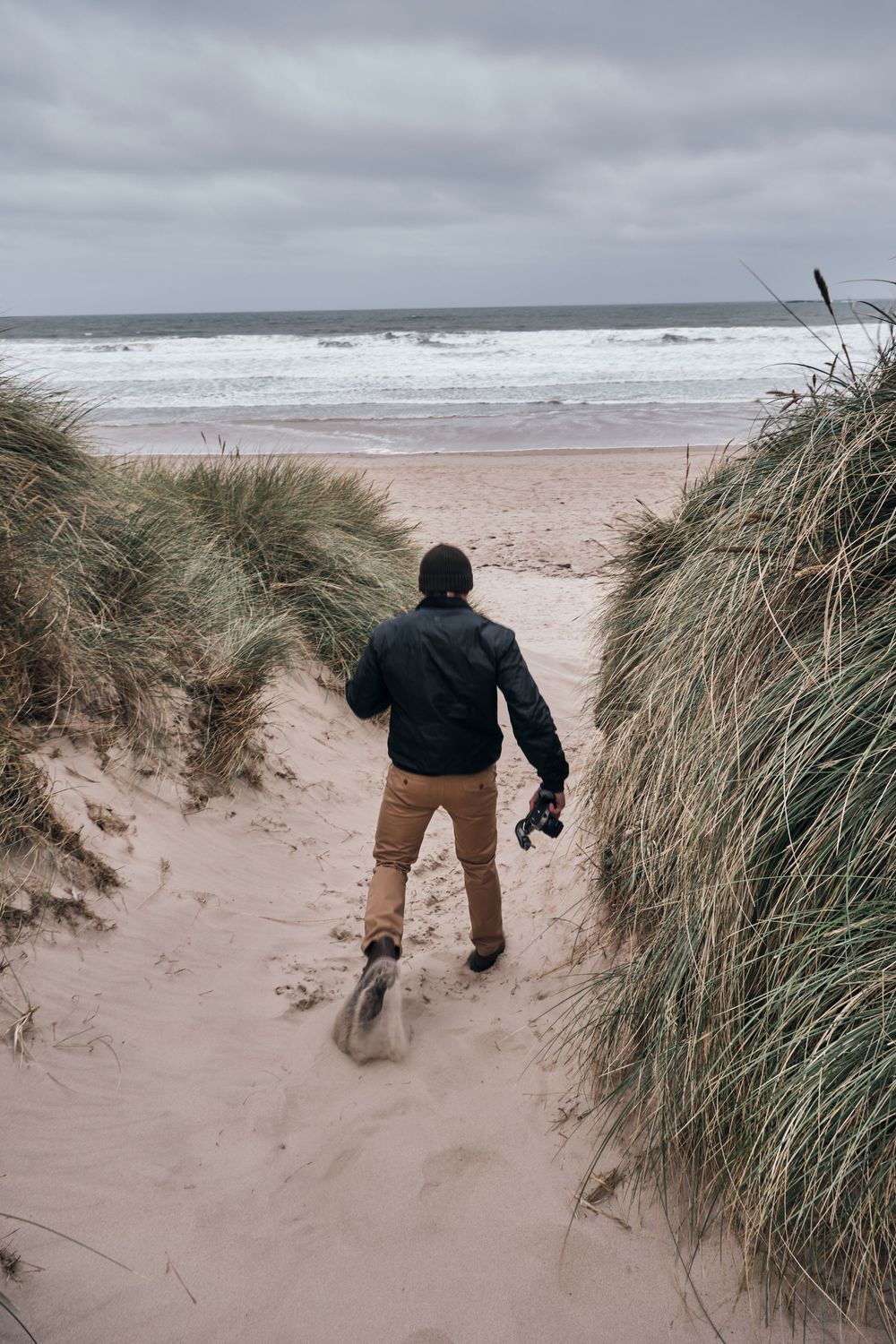 Photographer walking through sand dunes toward a stormy beach, holding camera, wearing black jacket and tan pants.