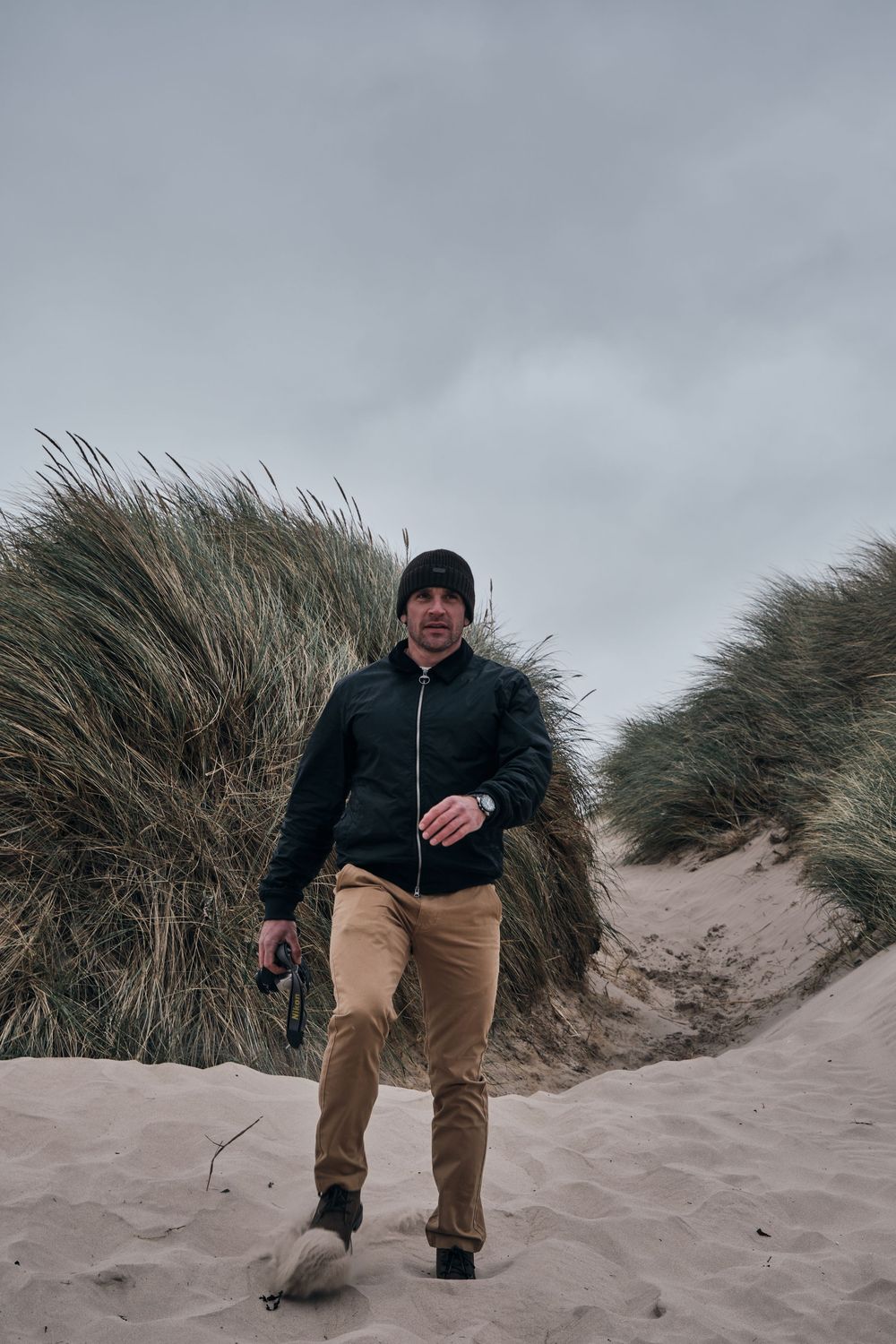 Man in black jacket and beanie walks through sandy dunes with tall grass on a cloudy day.