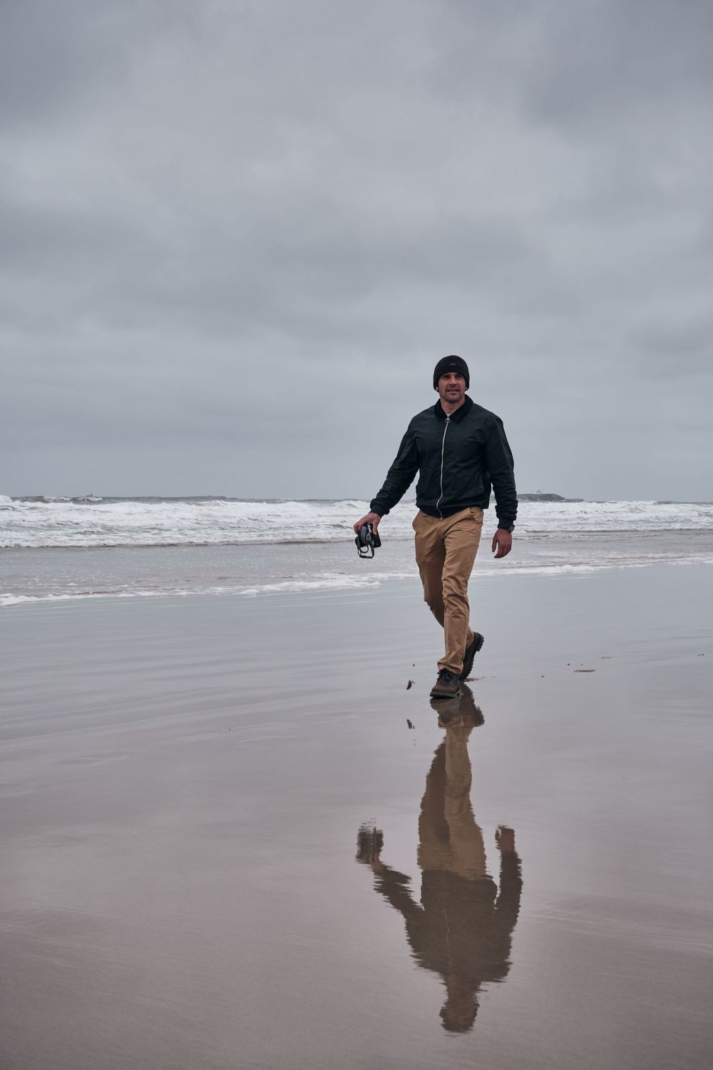 Man in dark jacket and beanie walks on a cloudy beach, his reflection visible on the wet sand near ocean waves.