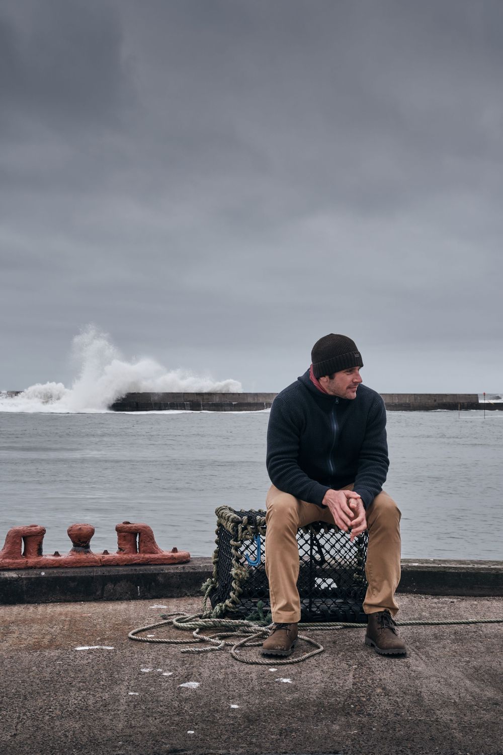 Man in black beanie and sweater sits on lobster trap at stormy harbor, waves crashing behind him.