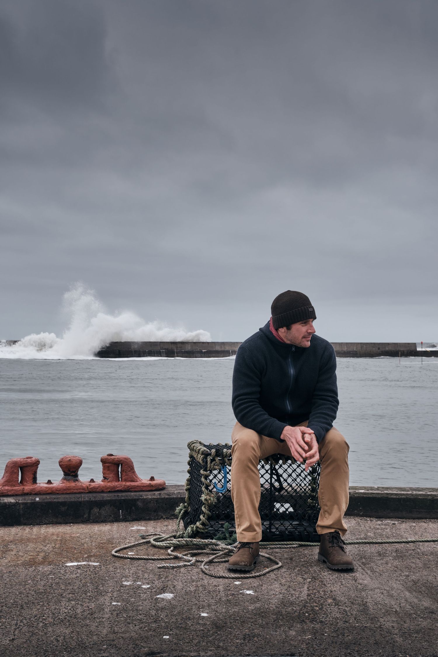 Man in black beanie and sweater sits on lobster trap at stormy harbor, waves crashing behind him.