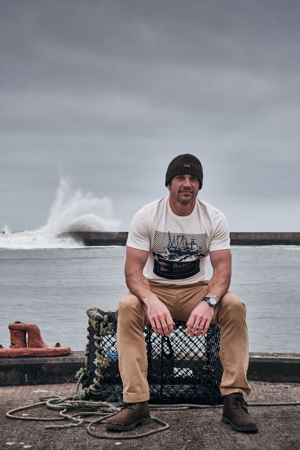 Man in white graphic tee and beanie sits on lobster trap at stormy harbor with crashing waves behind.