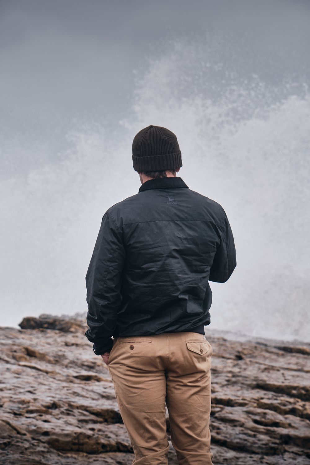 Man in black jacket and beanie stands on rocky shore watching powerful ocean waves crash behind him.