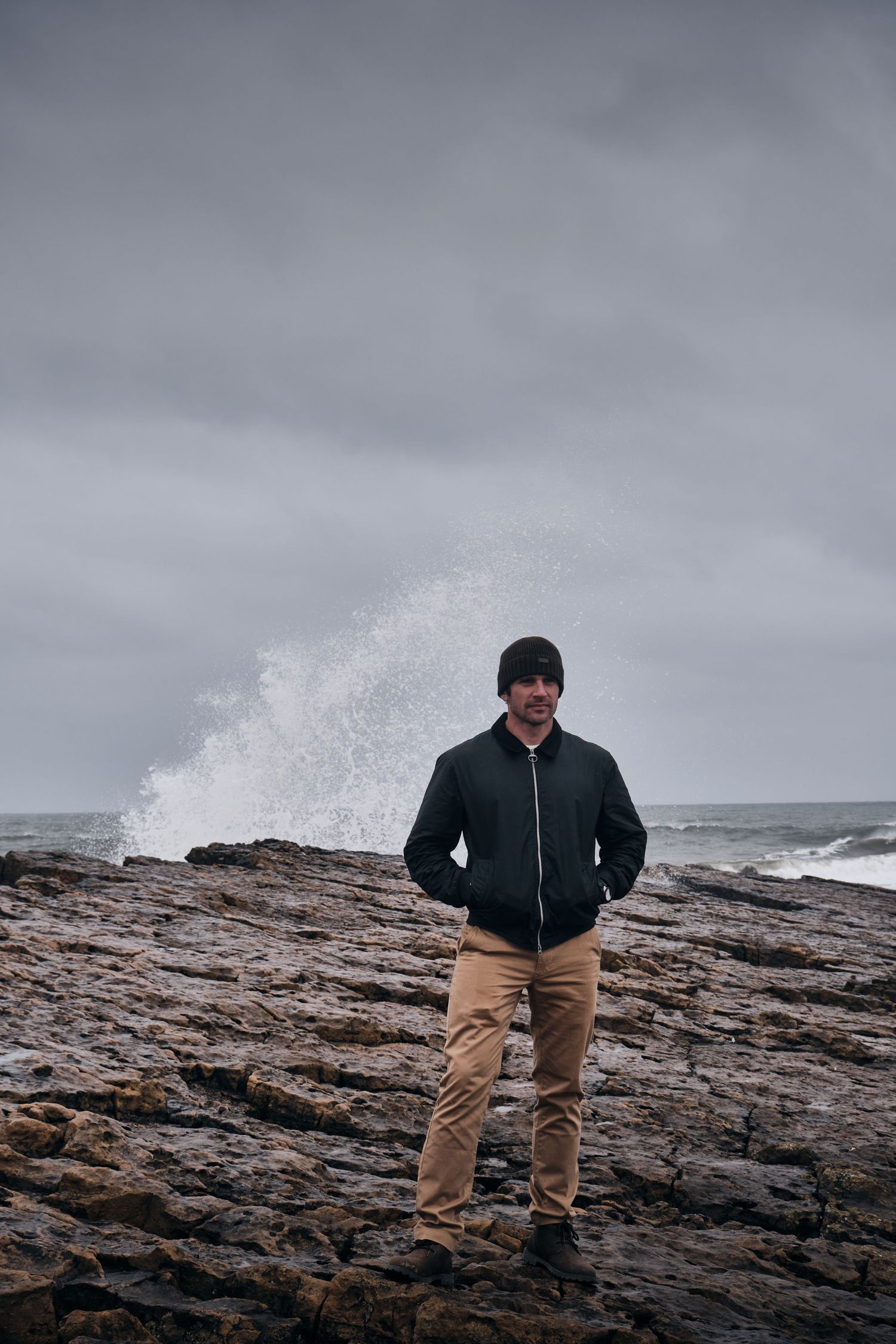 Man in dark jacket and beanie stands on rocky coastline with dramatic ocean waves crashing behind him.