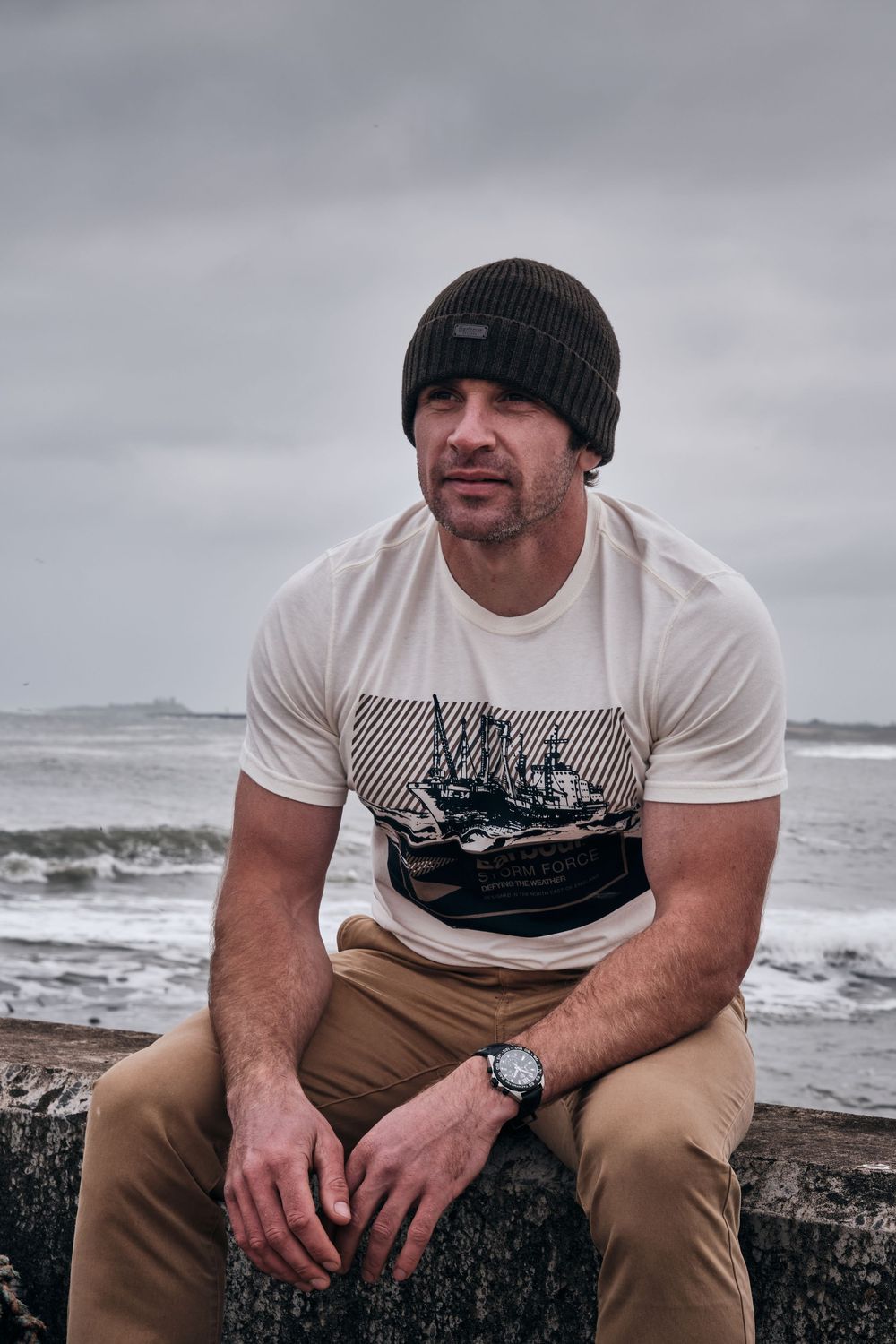Man in white graphic tee, khaki pants, and dark beanie sits on seaside wall with ocean waves behind him.