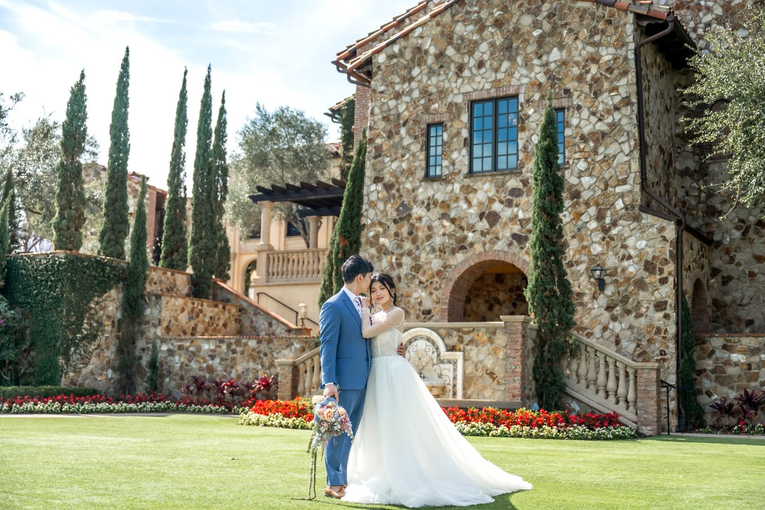 A romantic wedding photo at a luxurious Tuscan-style villa with stone walls and tall cypress trees in the background.