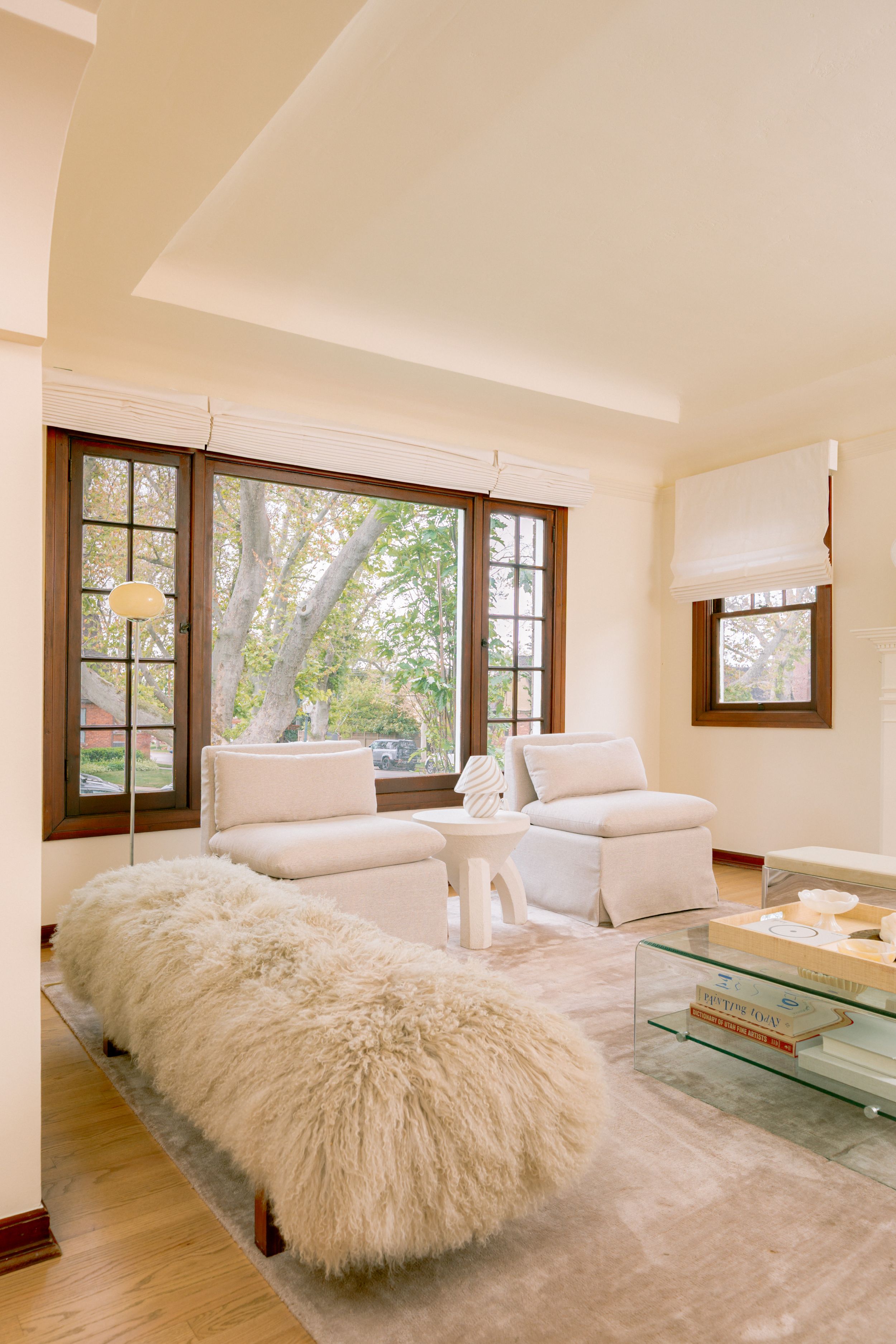 Bright living room with fluffy white bench, cream armchairs, glass coffee table, and large wood-framed windows overlooking trees.