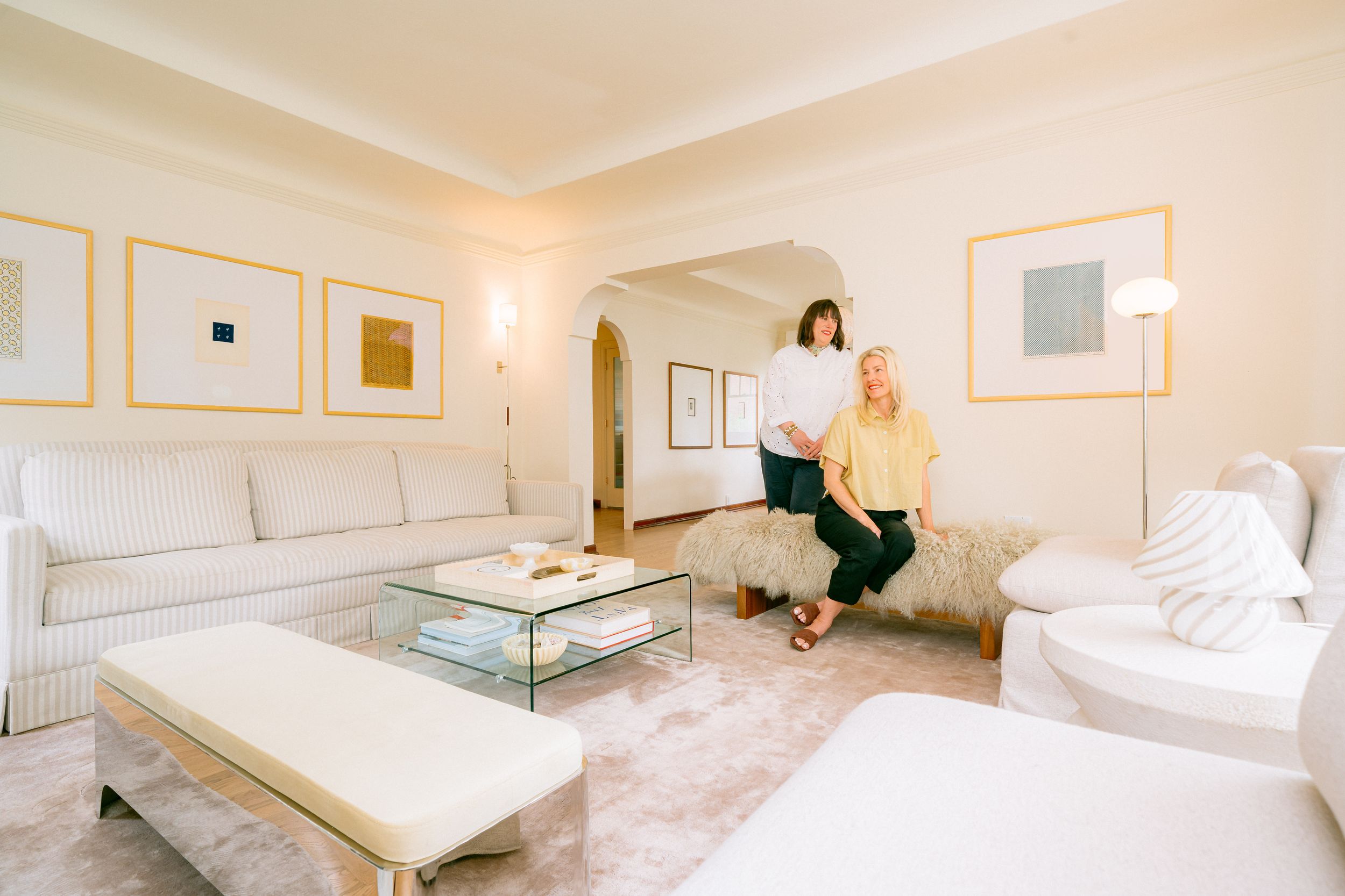 Two women sitting in an elegant white living room with gold-framed artwork and classic furniture.