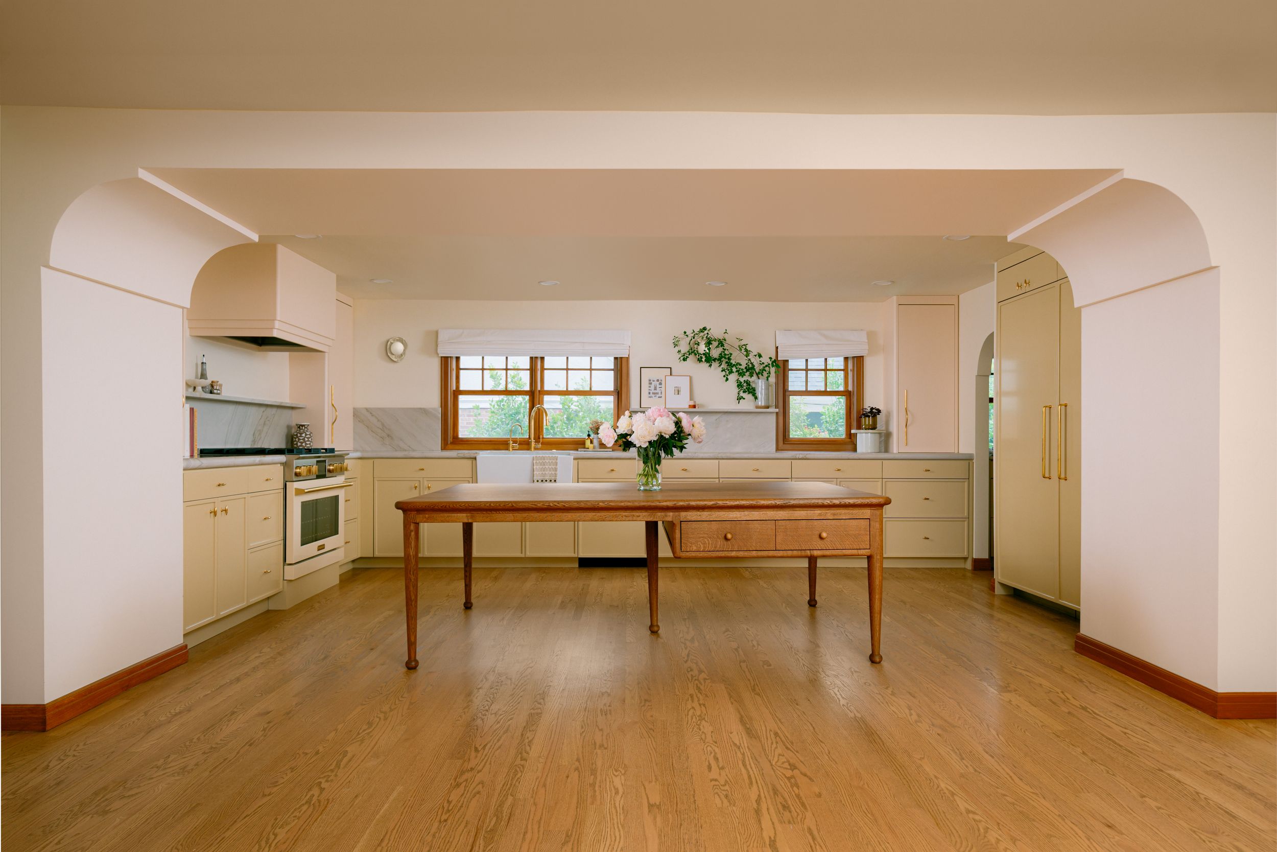 Bright kitchen with hardwood floors, arched doorways, wooden dining table, light cabinets, and windows with fresh flowers.
