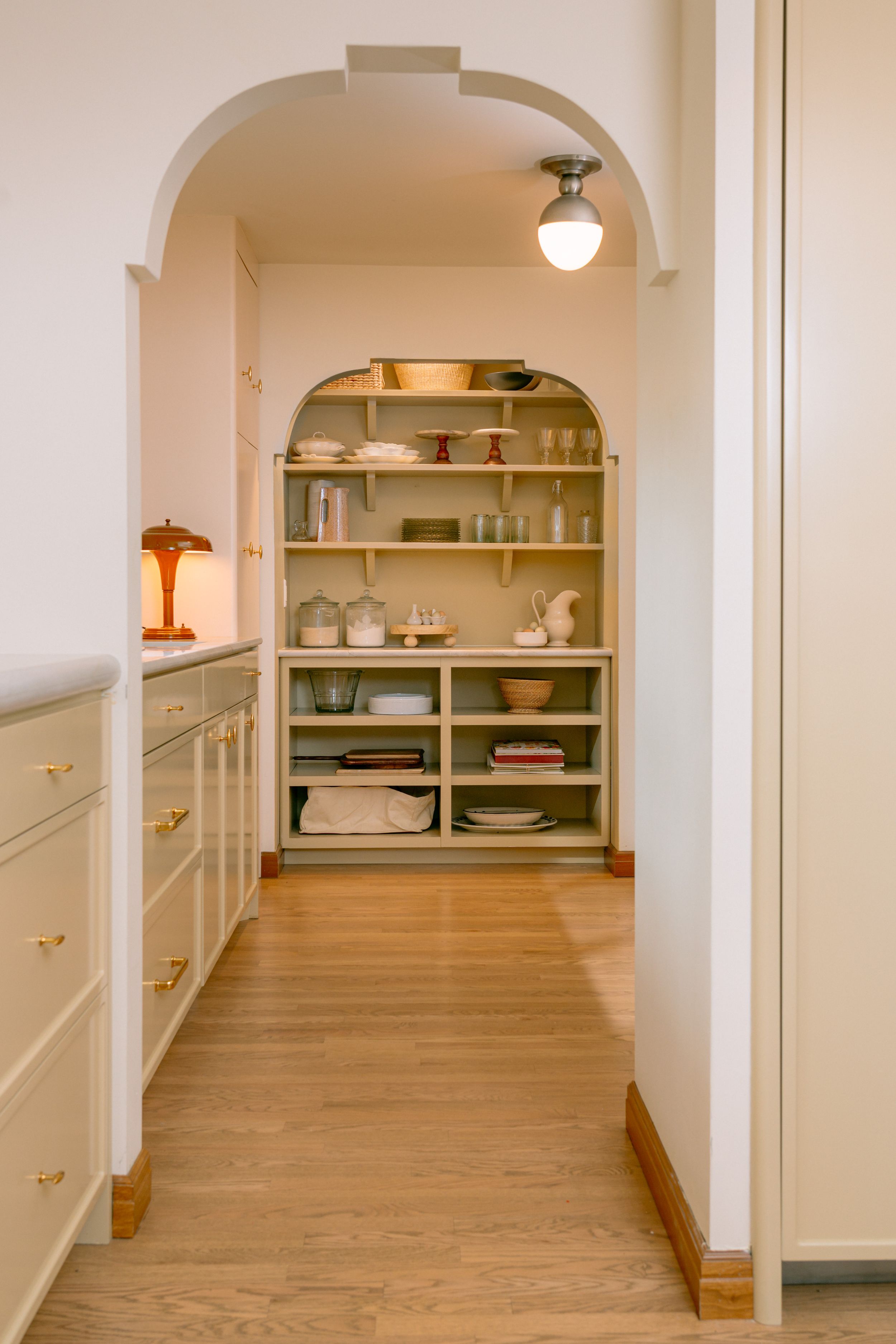 Elegant butler pantry with arched doorway, sage green shelving, brass hardware cabinets, and light wood floors.
