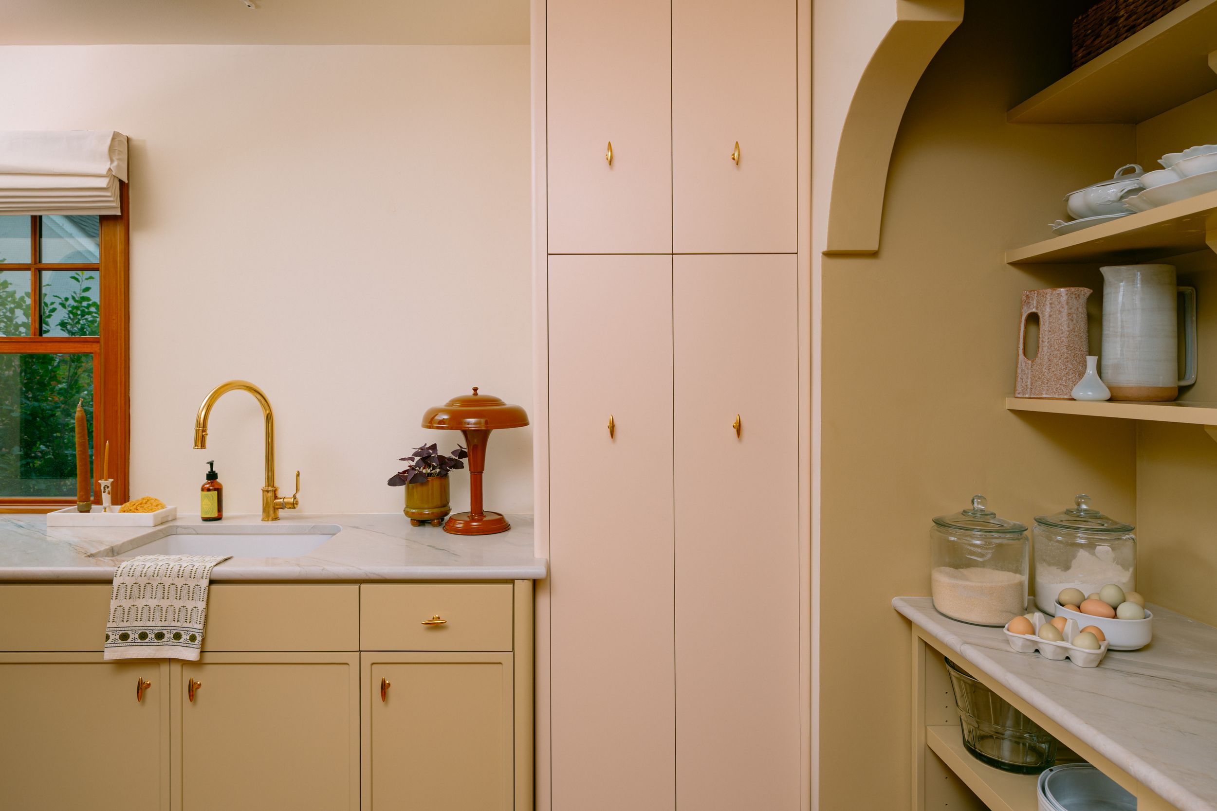 Bright kitchen with blush pink cabinets, gold faucet, marble countertop, and open shelving pantry displaying jars and bowls.