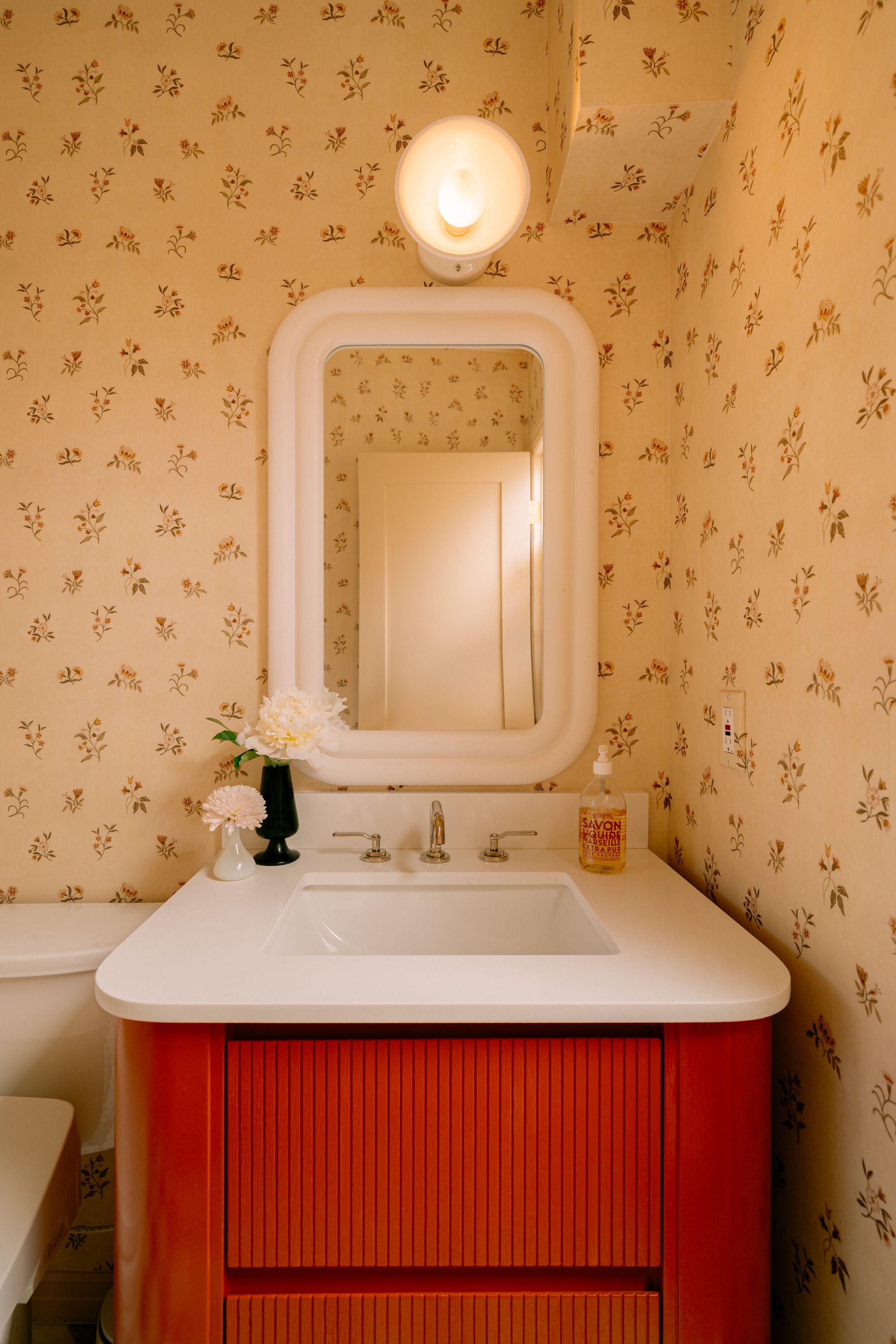 Vintage bathroom with floral wallpaper, red vanity, white countertop, framed mirror, and wall sconce light fixture.