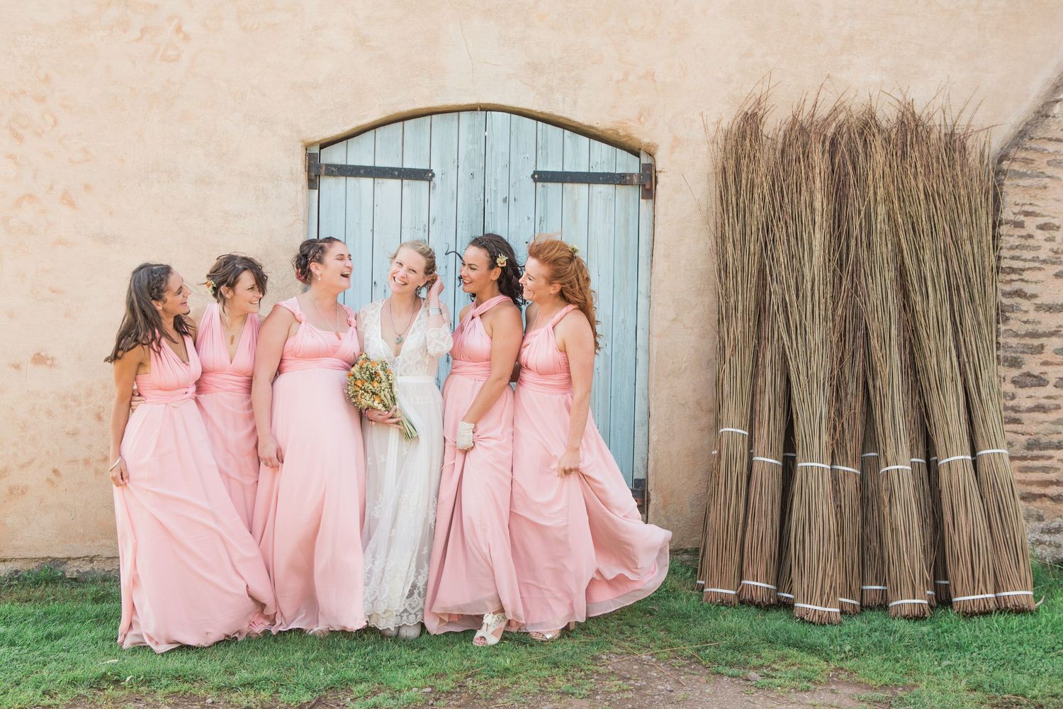 Bride and bridesmaids in pink gowns laughing together near rustic blue door with bundled brooms at outdoor wedding venue.
